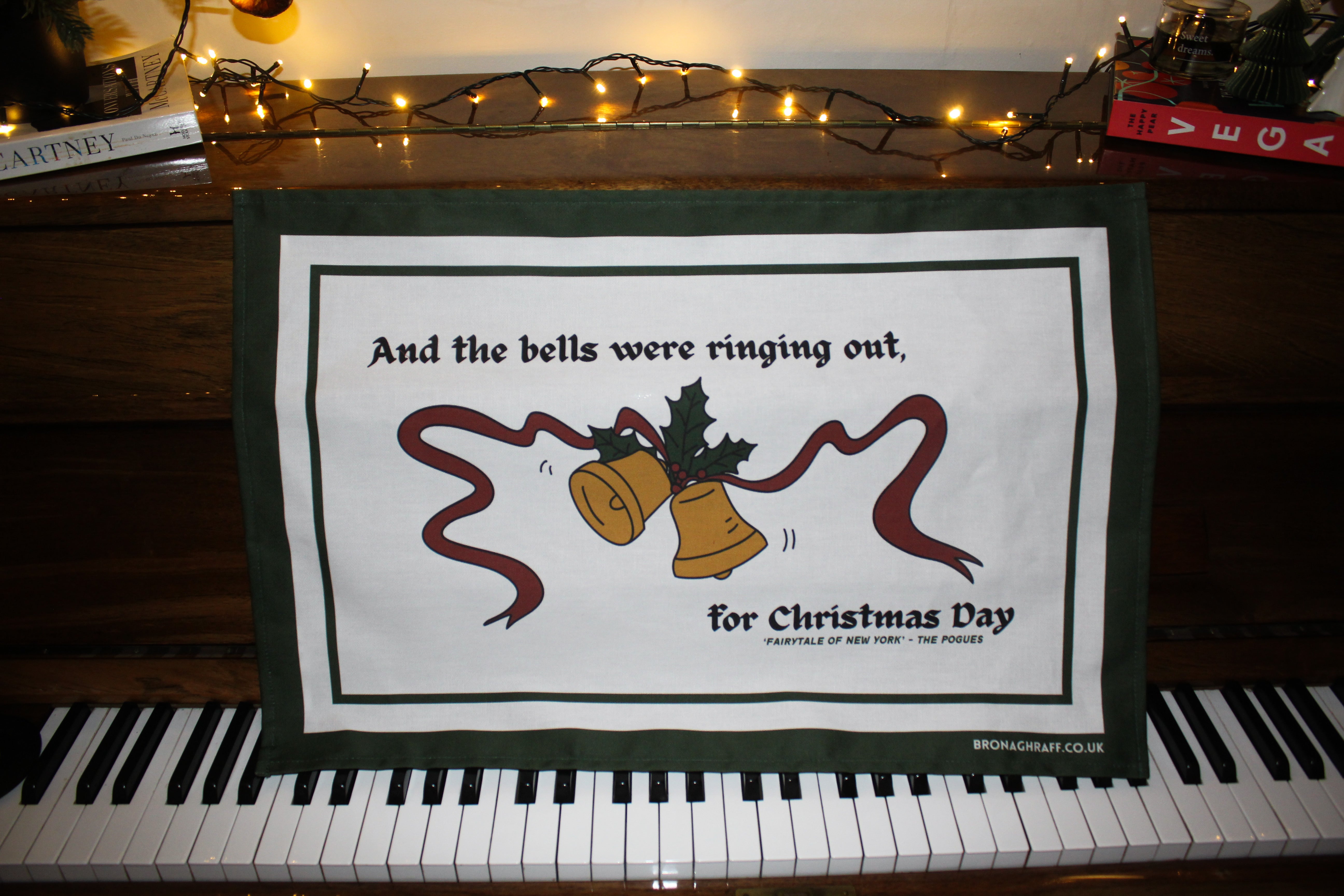 festive Irish tea towel with the quote 'And the bells were ringing out' from Fairytale of New York, featuring golden bells and holly on organic cotton.