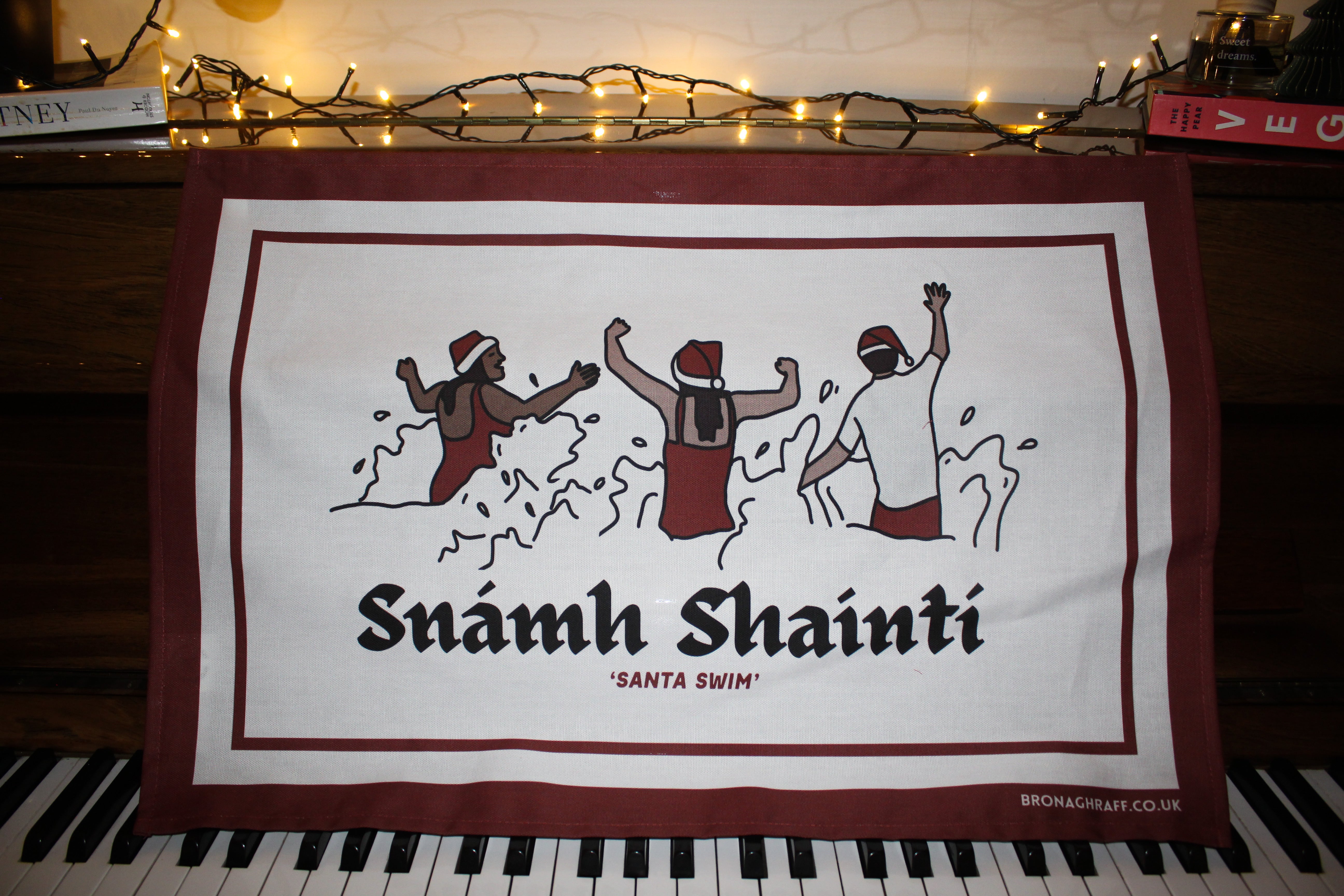 An Irish Christmas tea towel featuring swimmers in festive hats with the phrase 'Snámh Shainti,' celebrating the Christmas Day swim tradition