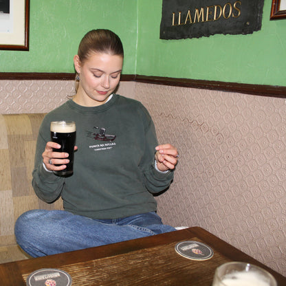 Woman sitting at a bar holding a pint of guinness, wearing and irish christmas jumper with a green wall and framed pictures in the background.
