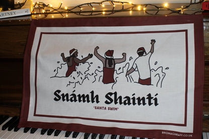 Organic cotton tea towel with Irish text 'Snámh Shainti' and festive illustrations of swimmers enjoying a Christmas swim